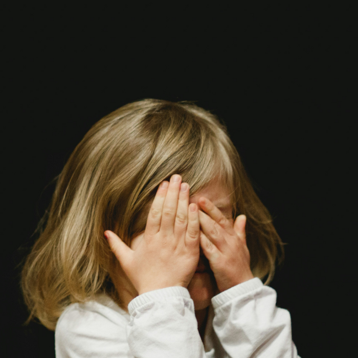 young girl with blonde hair holding her hands over her face. Background is black.