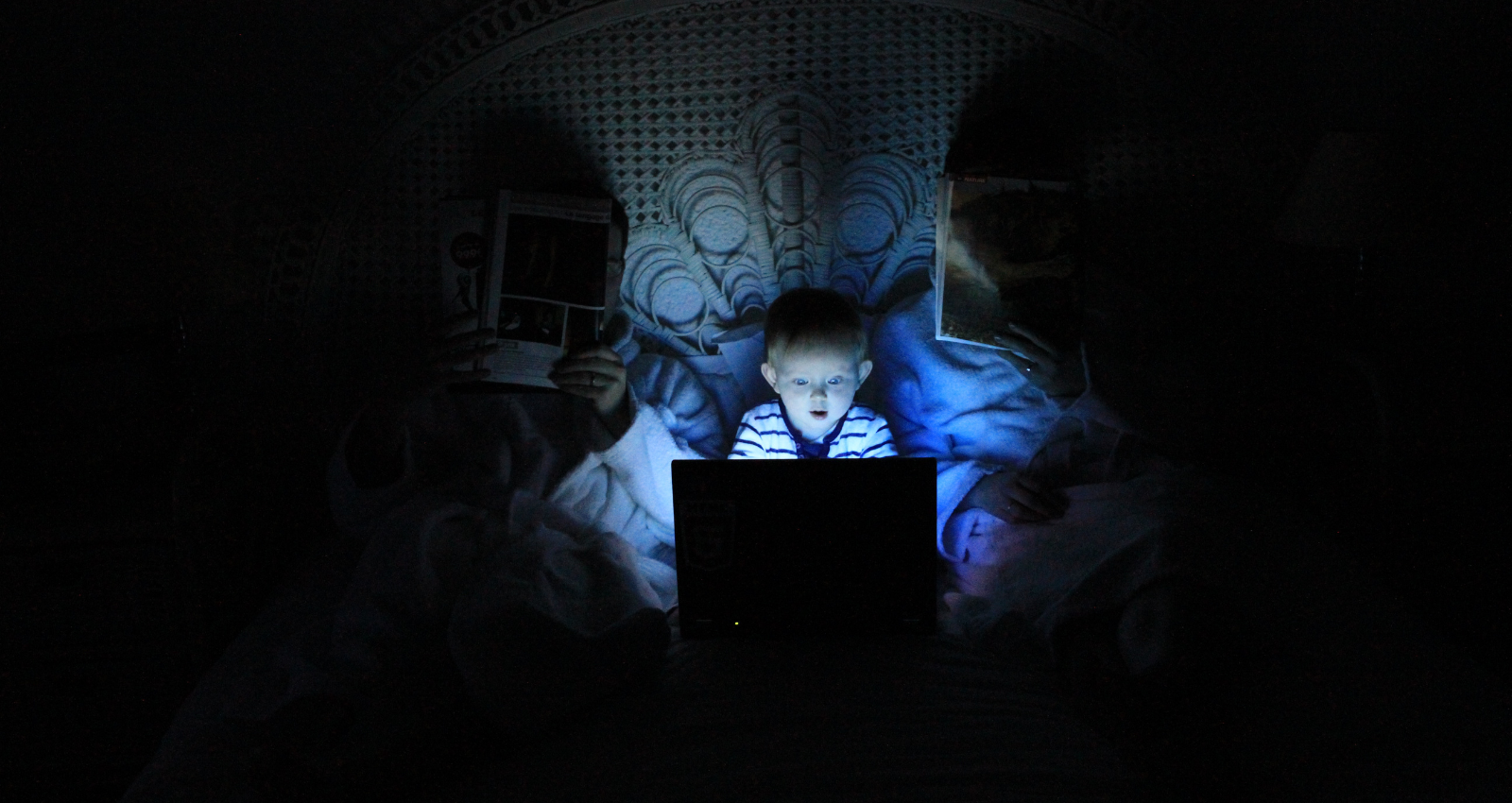 Internet Safety. A baby sitting in between parents in bed, the room is pitch black. A light is shining on his face from looking at a computer screen.