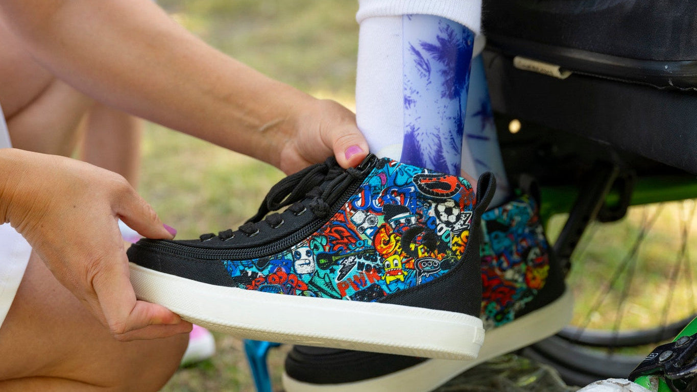 A close-up lifestyle shot of a person holding a black low-top sneaker with a vibrant multicolor floral and skull graphic pattern on the side, while another person sits in a wheelchair nearby.